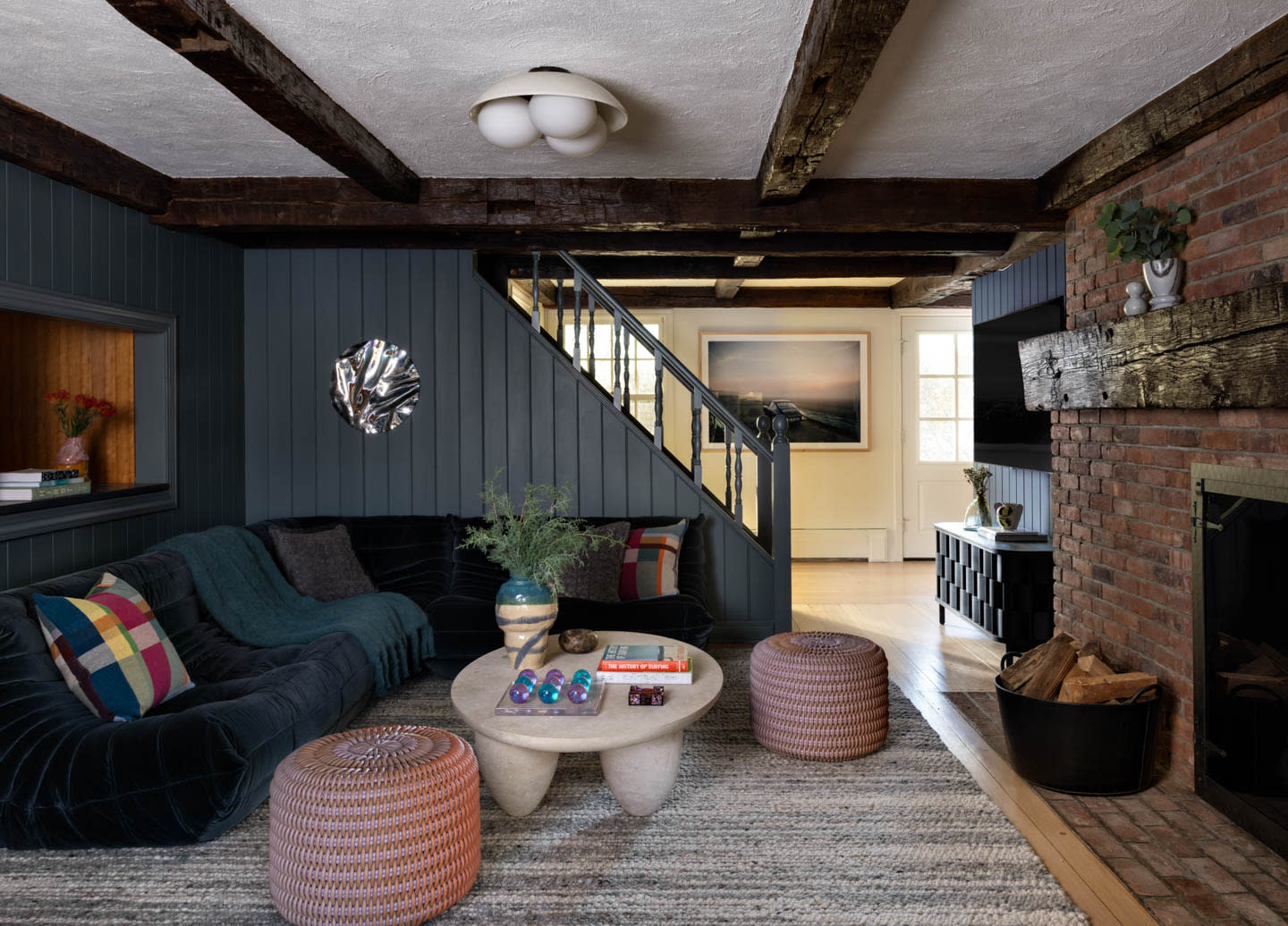 Cozy living room with rustic beams, dark paneled walls, and brick fireplace. Features a velvet sectional, round table, woven poufs, and colorful cushions.