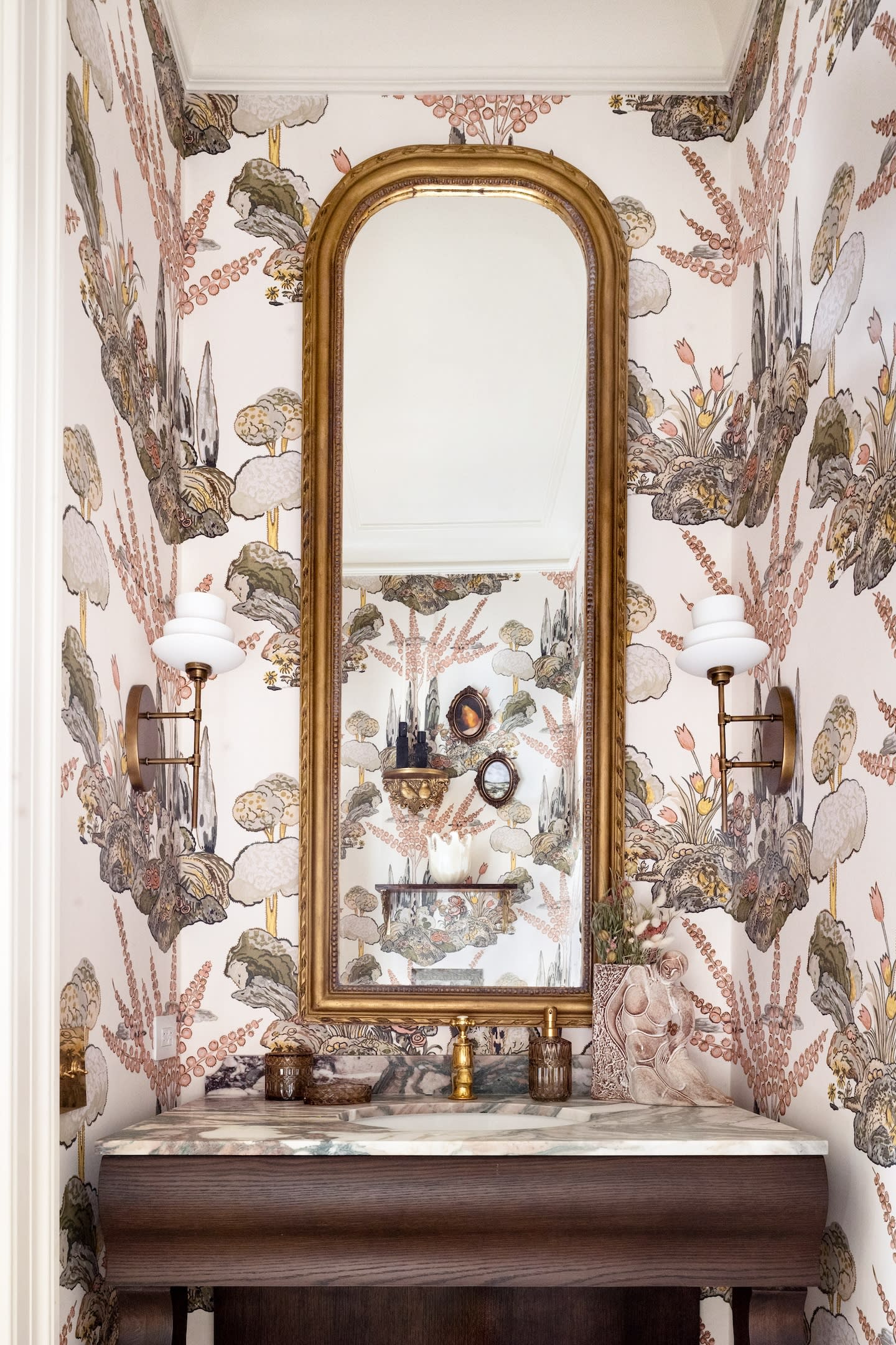 Luxurious restroom with botanical wallpaper, a marble sink, and a large ornate mirror. Brass sconces and elegant decor create a sophisticated atmosphere.