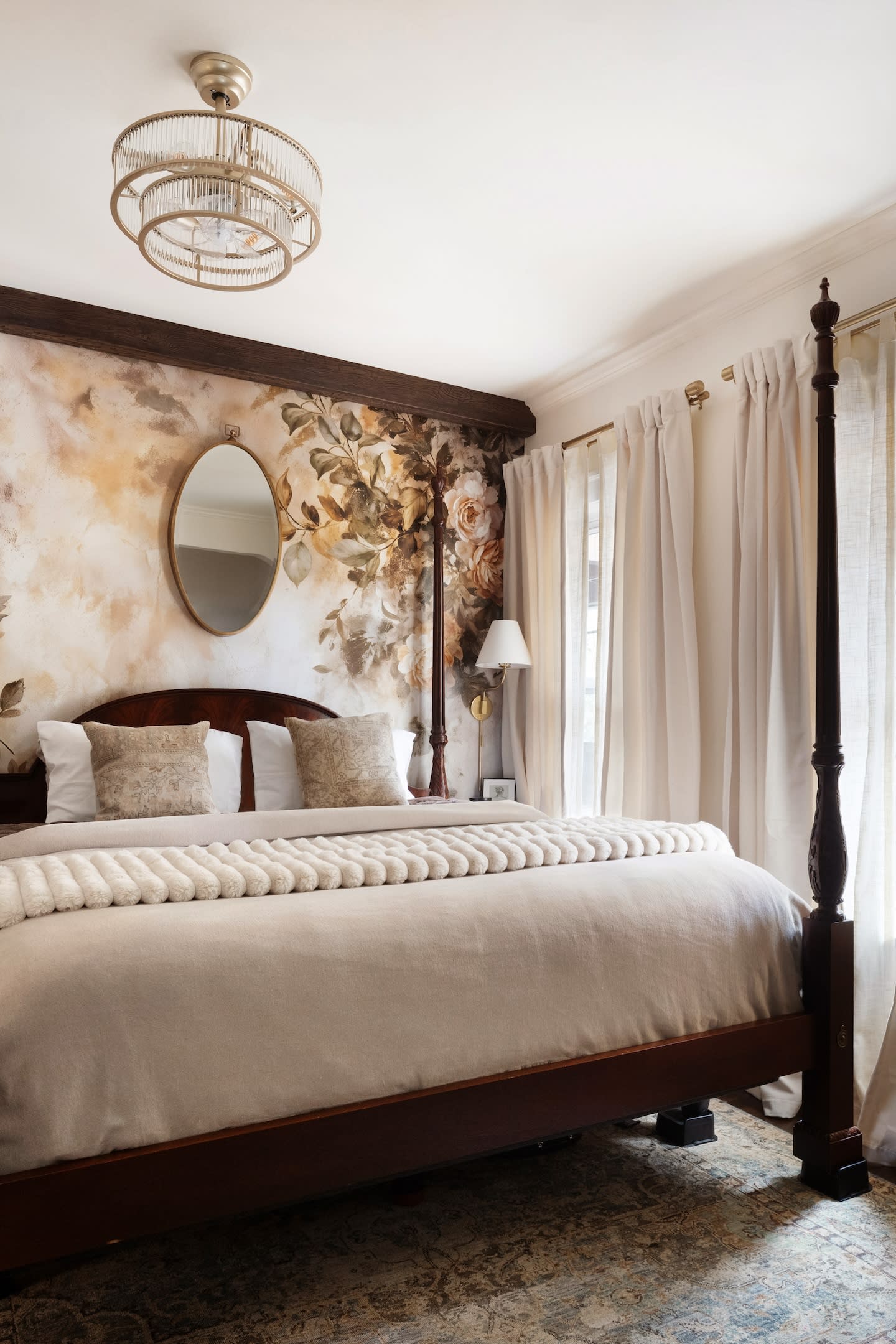 Elegant bedroom with a four-poster bed, floral accent wall, oval mirror, and soft curtains. Warm tones create a cozy, inviting atmosphere.