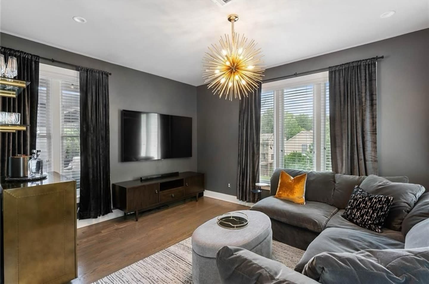 Modern living room with dark gray walls, a large L-shaped sofa, and a stylish starburst light fixture. Large windows with black curtains add elegance.